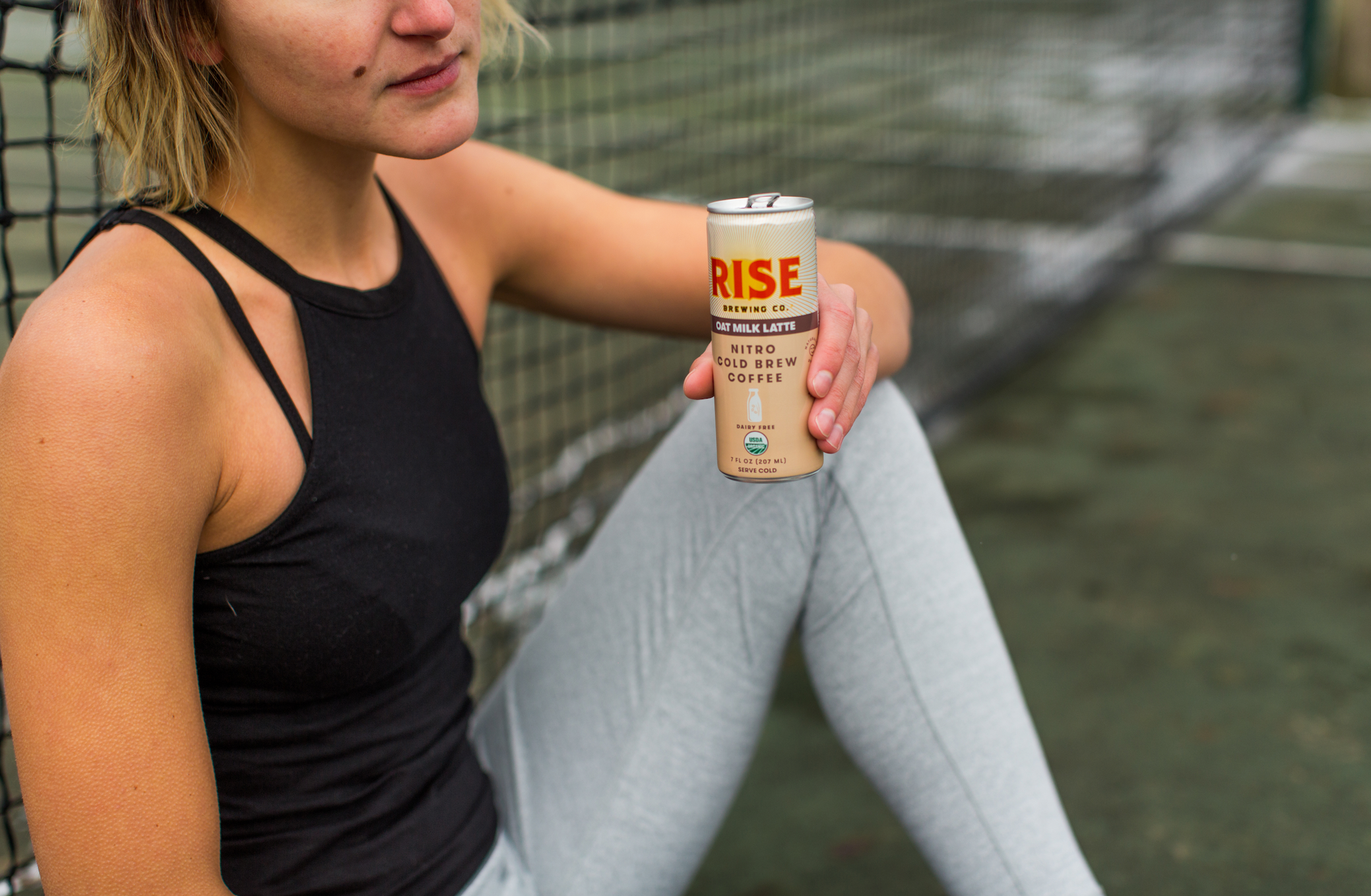 Sporty woman on tennis court with RISE Nitro Brewing Co. Oat Milk Latte can in her hand