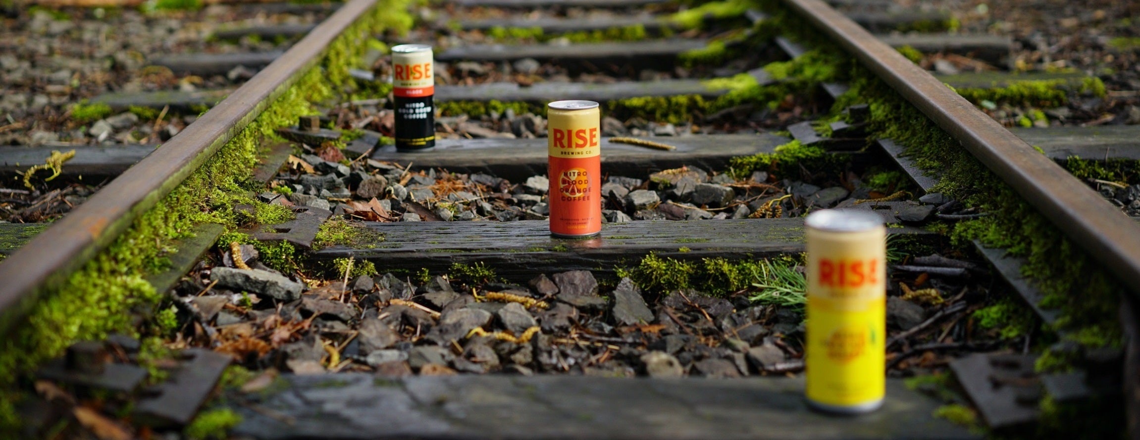 Cans of RISE Coffee on train tracks - Original Black, Blood Orange, and Lemonade flavors of nitro cold brew coffee